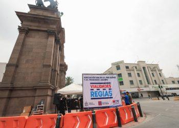 Large stone monument on the left with an urban roadwork setup: orange barriers in the foreground, a white banner reading 'VIALIDADES RÉGIAS' and a tent nearby. A few people are visible around the area.