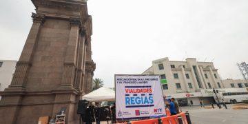 Large stone monument on the left with an urban roadwork setup: orange barriers in the foreground, a white banner reading 'VIALIDADES RÉGIAS' and a tent nearby. A few people are visible around the area.