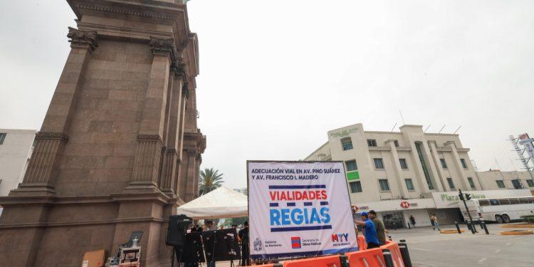 Large stone monument on the left with an urban roadwork setup: orange barriers in the foreground, a white banner reading 'VIALIDADES RÉGIAS' and a tent nearby. A few people are visible around the area.