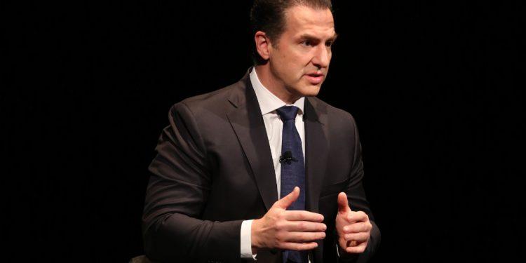 Man in a dark suit and blue tie sits on a stage, speaking into a lapel microphone.