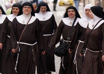 Group of nuns walking together down a city street in brown habits with white collars and veils, some carrying bags and rosaries attached to belts.