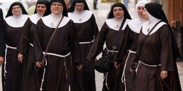 Group of nuns walking together down a city street in brown habits with white collars and veils, some carrying bags and rosaries attached to belts.