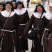 Group of nuns walking together down a city street in brown habits with white collars and veils, some carrying bags and rosaries attached to belts.