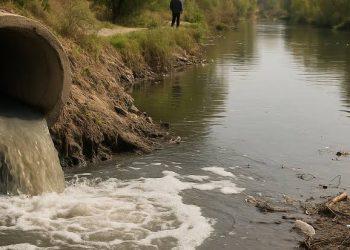 Concrete pipe discharging murky water into a canal; a person stands in the background on the bank.