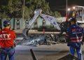 Nighttime street scene of rescue workers watching an excavator clear rubble from a broken road, MTY shirts visible.