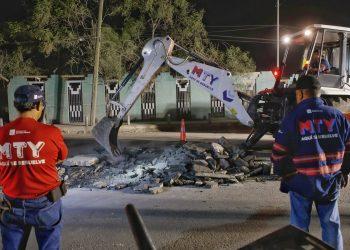 Nighttime street scene of rescue workers watching an excavator clear rubble from a broken road, MTY shirts visible.