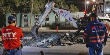 Nighttime street scene of rescue workers watching an excavator clear rubble from a broken road, MTY shirts visible.