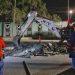 Nighttime street scene of rescue workers watching an excavator clear rubble from a broken road, MTY shirts visible.